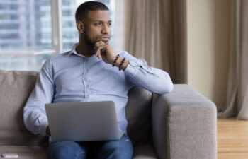 A man sits on a couch with a laptop on his lap, resting his chin on his hand and looking thoughtfully out a window.