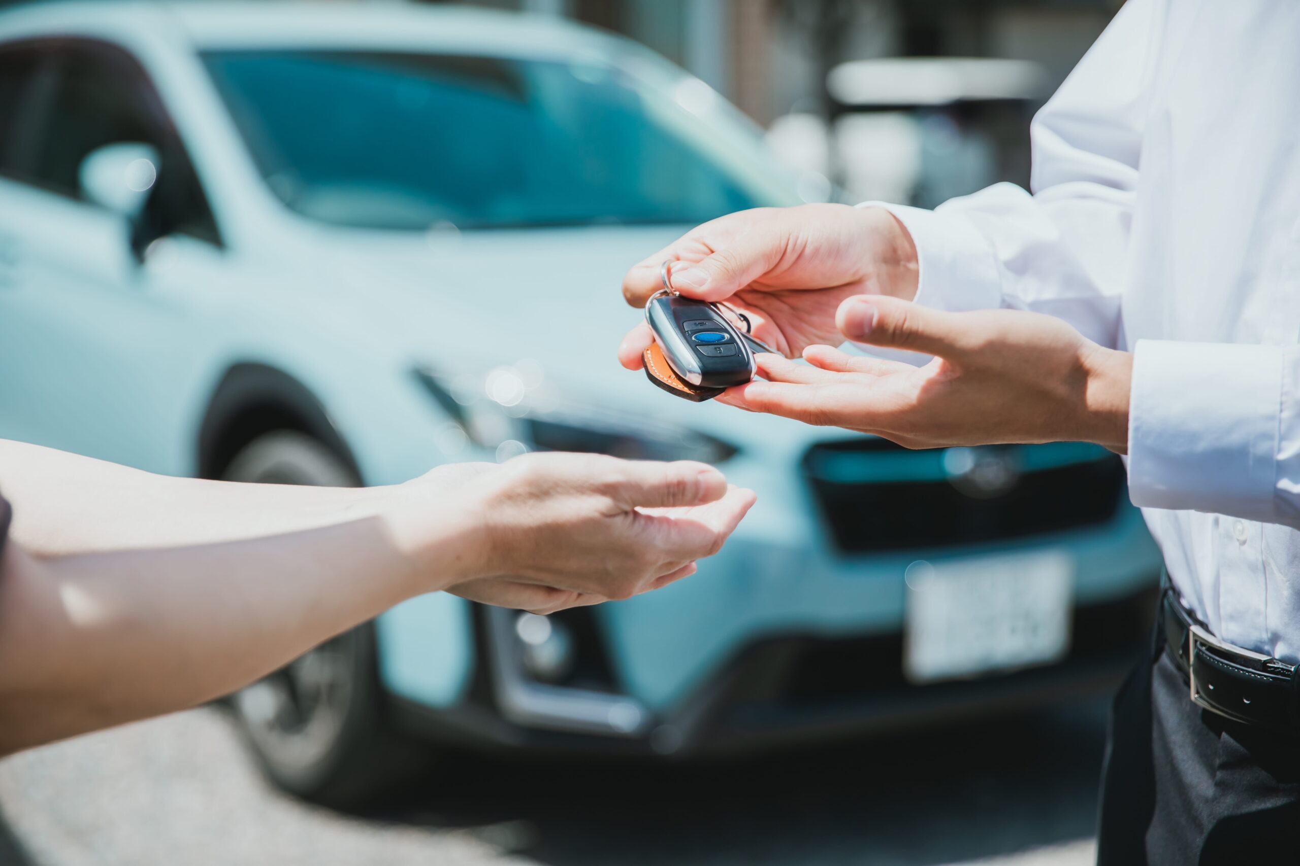 A person hands car keys to another person in front of a light blue car parked outdoors.