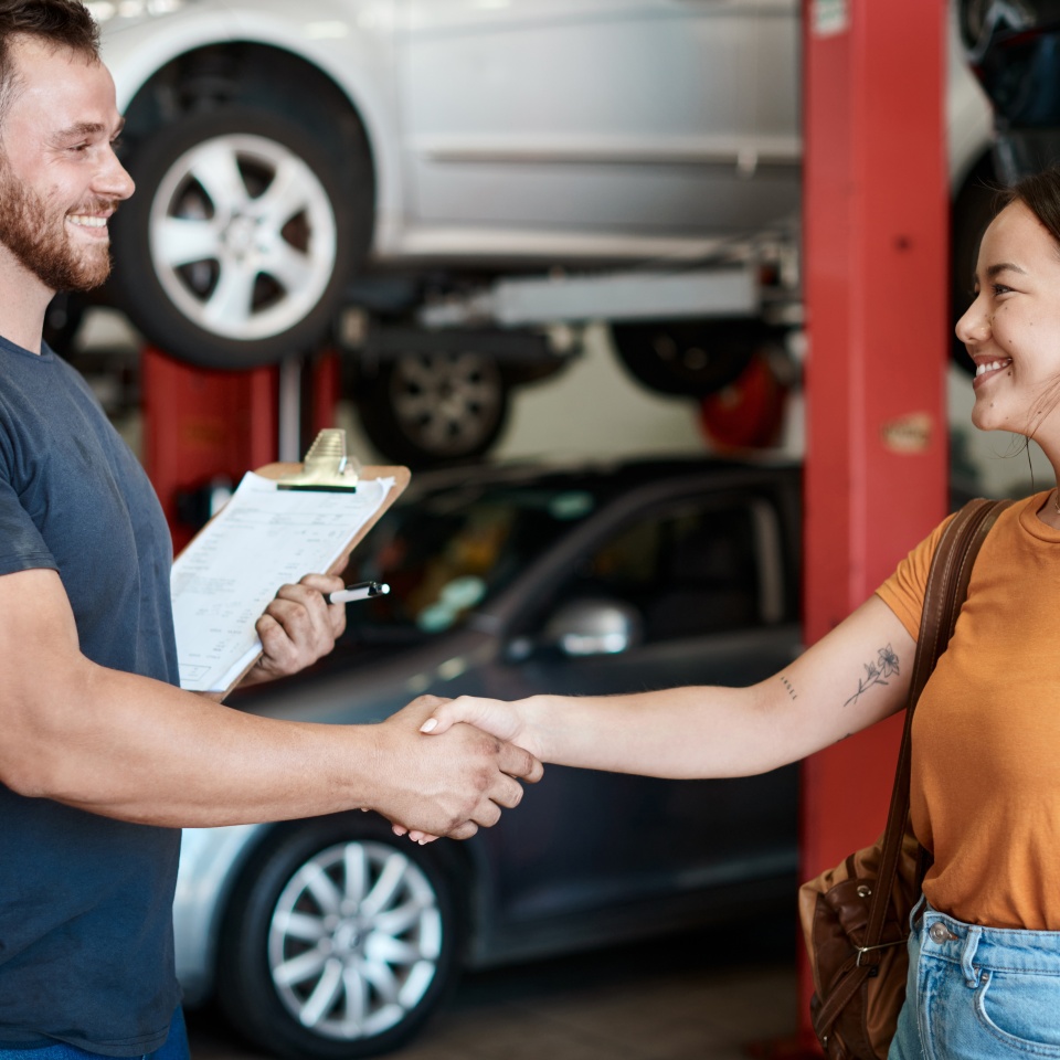 Shaking hands, smile and woman with mechanic in workshop for deal on vehicle repair or maintenance. Clipboard, welcome and Asian person with industrial worker for handshake in garage for car service.