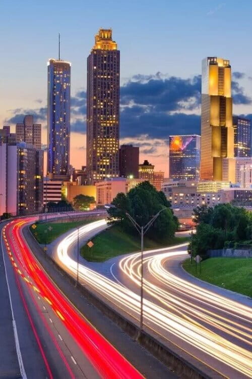 Atlanta city skyline at sunset with light trails from traffic on a highway in the foreground and modern buildings in the background.
